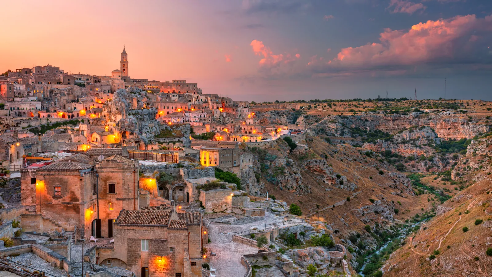 The Sassi di Matera at dusk, with lit stone houses, a church and a bell tower beside the Gravina gorge.