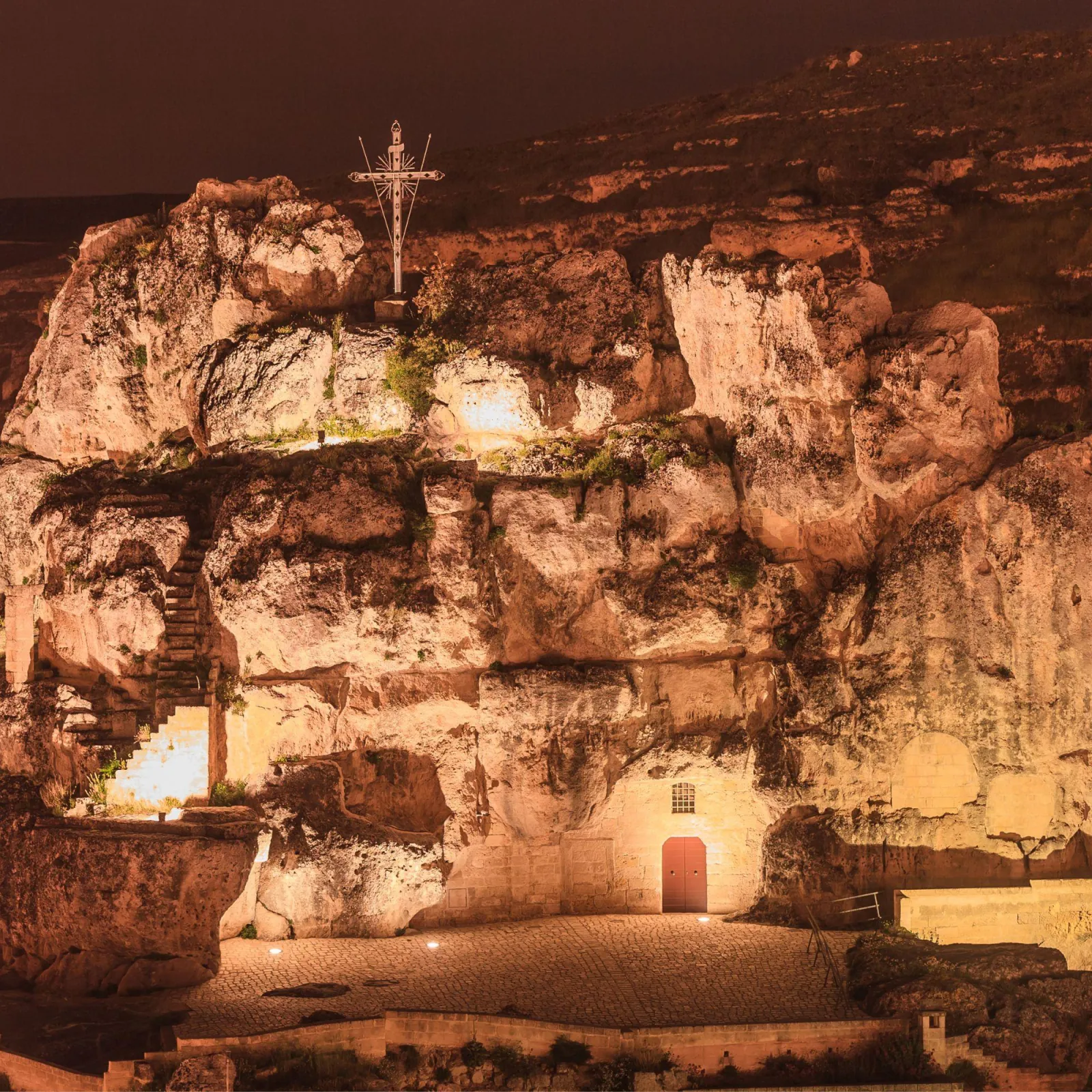 A rock-cut church complex in Matera lit up at night, with a cross on the limestone crag above.