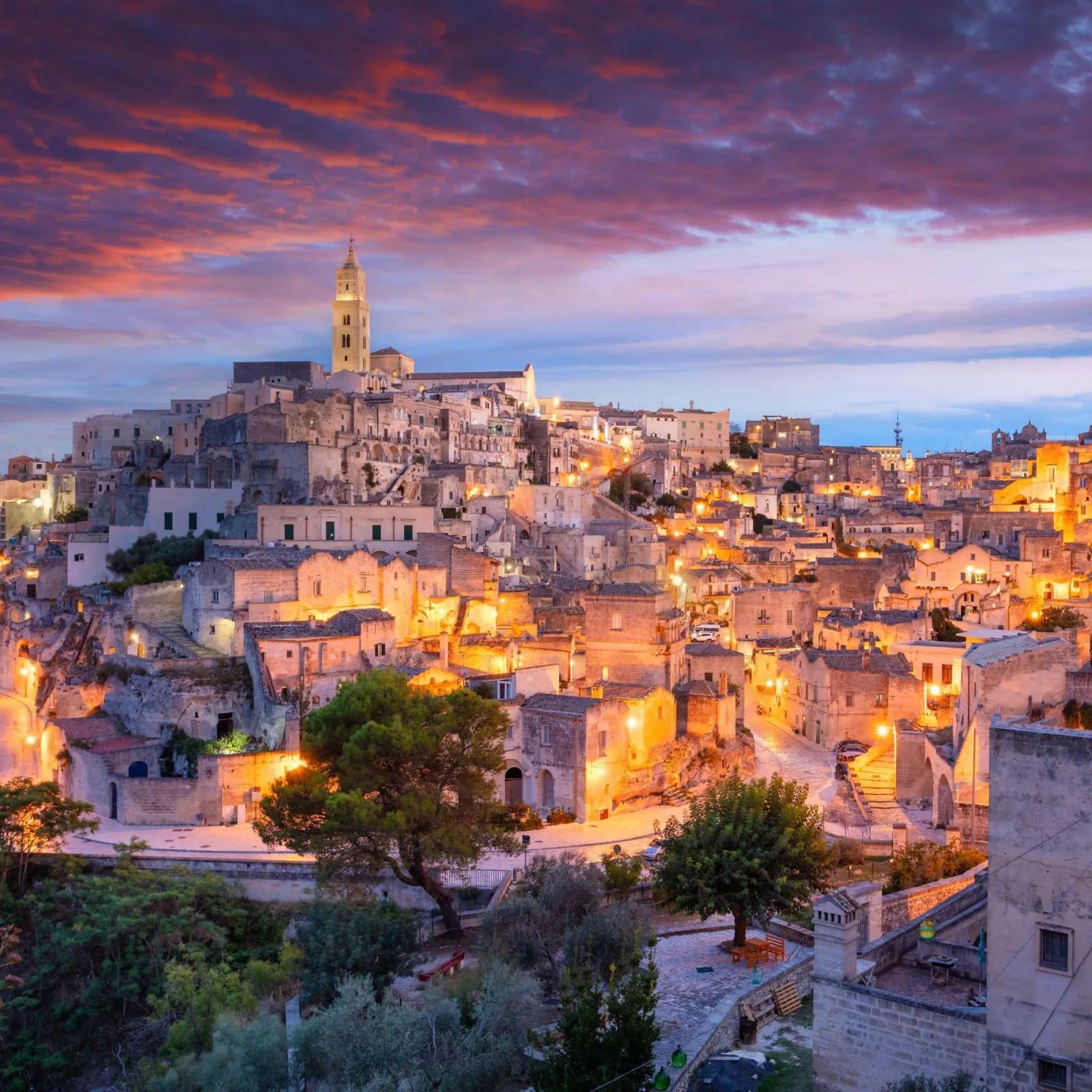 Matera's old town glowing at dusk under a purple and orange sky, the cathedral bell tower above the roofs.