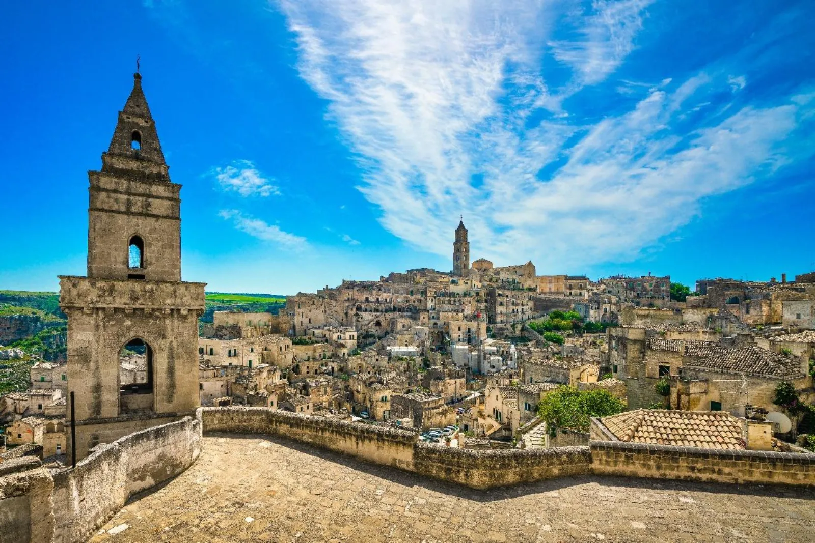 A stone bell tower on a terrace overlooks the Sassi di Matera under a bright blue sky with wispy cloud.