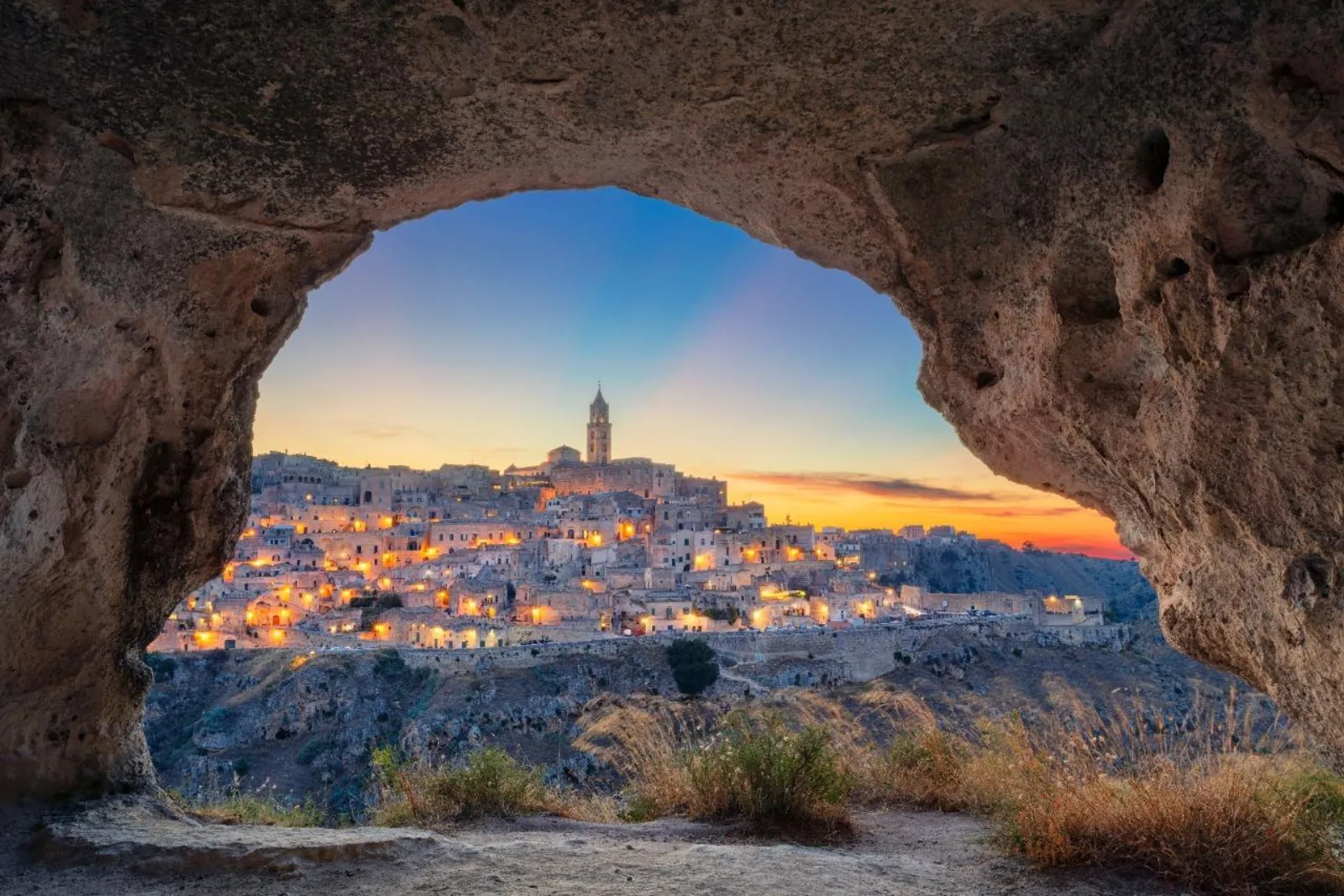 The Sassi of Matera glowing at dusk, seen through a natural rock arch in the cliff.