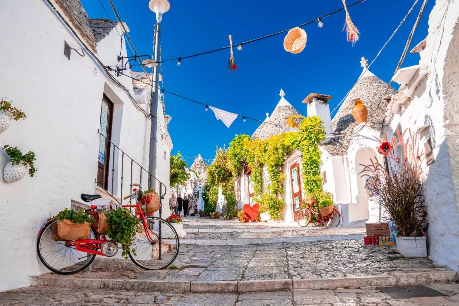 A red bicycle with flower baskets leans on a whitewashed wall in a trulli lane in Alberobello.