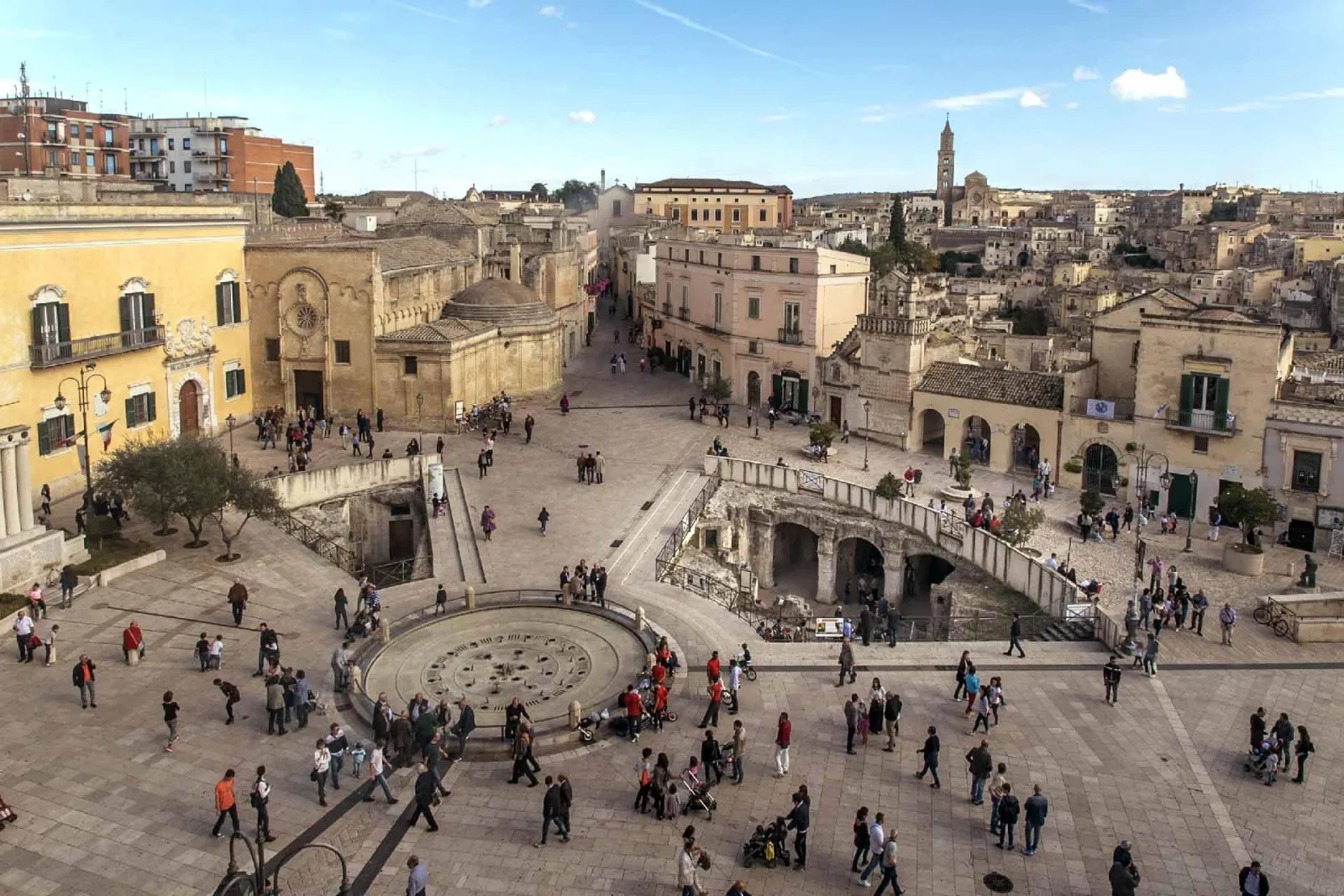 A busy stone piazza with a round fountain and arcades seen from above, with a bell tower behind.