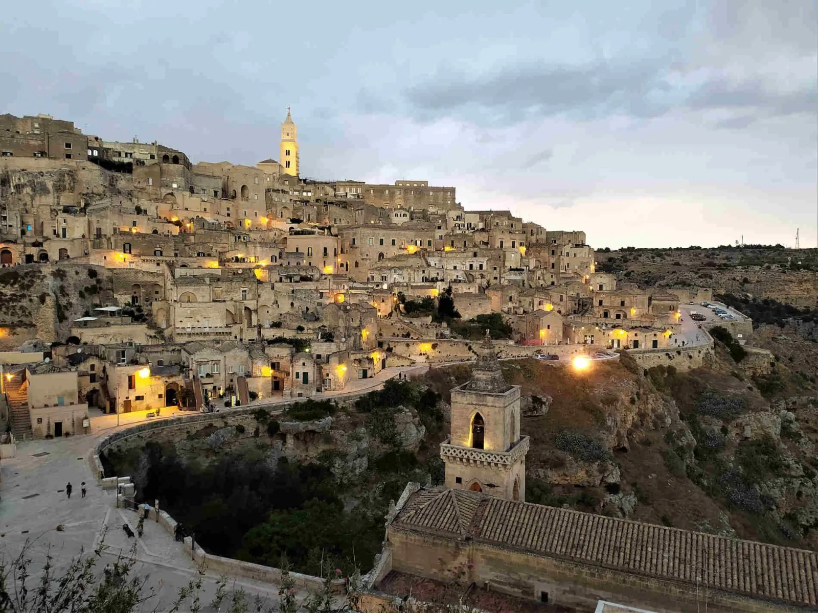 Matera at dusk, with the stone houses of the Sassi lit up and a small bell tower above the gorge.