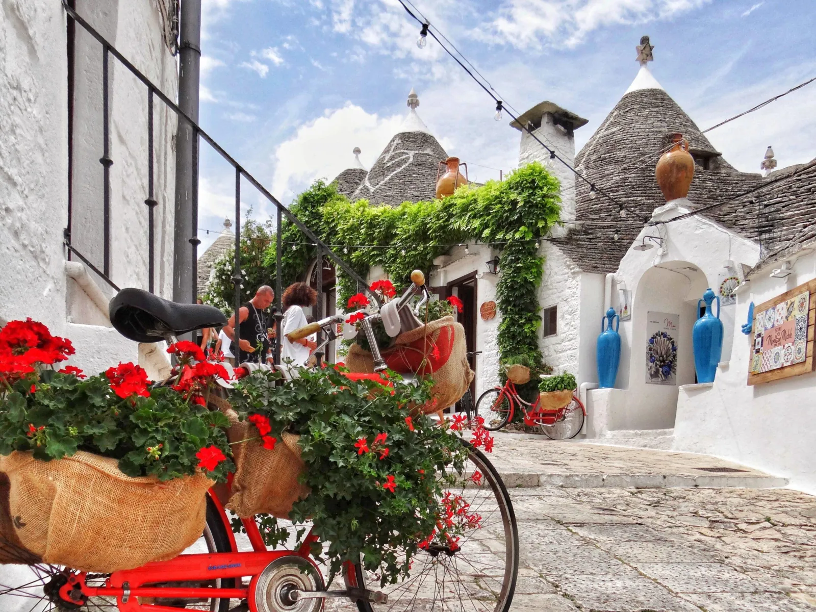 A red bicycle with a basket of red flowers stands before whitewashed trulli with grey conical roofs in Alberobello.
