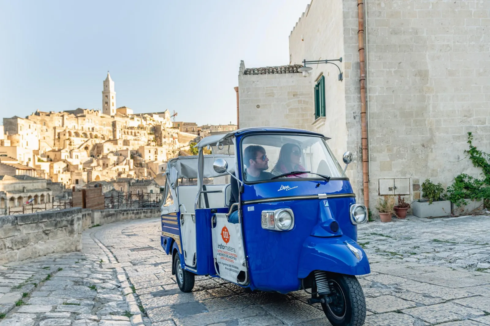 A blue and white Ape Calessino tuk-tuk on a cobbled bend, with the Sassi and cathedral tower behind.