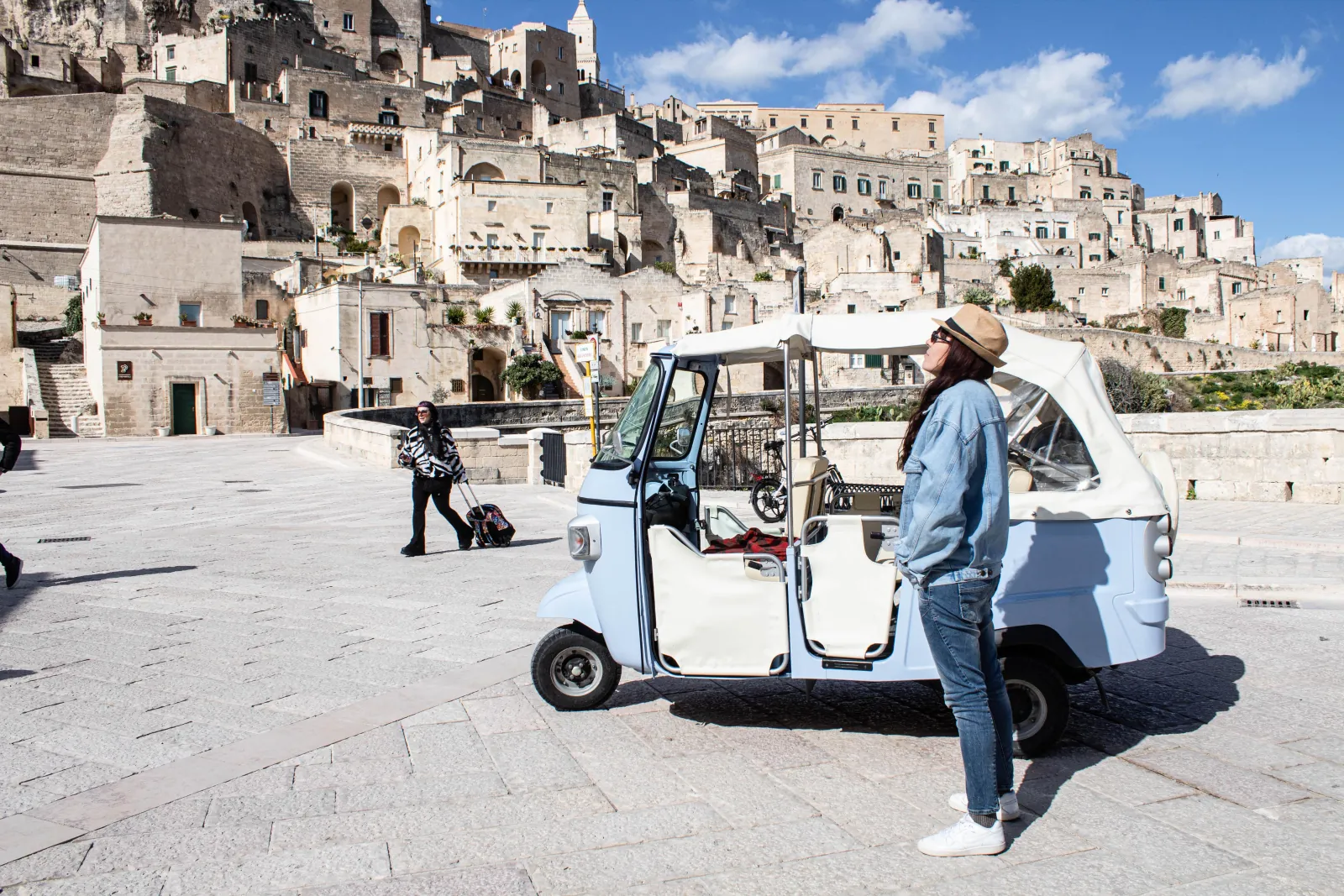 A woman in a sun hat stands beside a pale blue Ape tuk-tuk on a Matera square, Sassi houses behind.