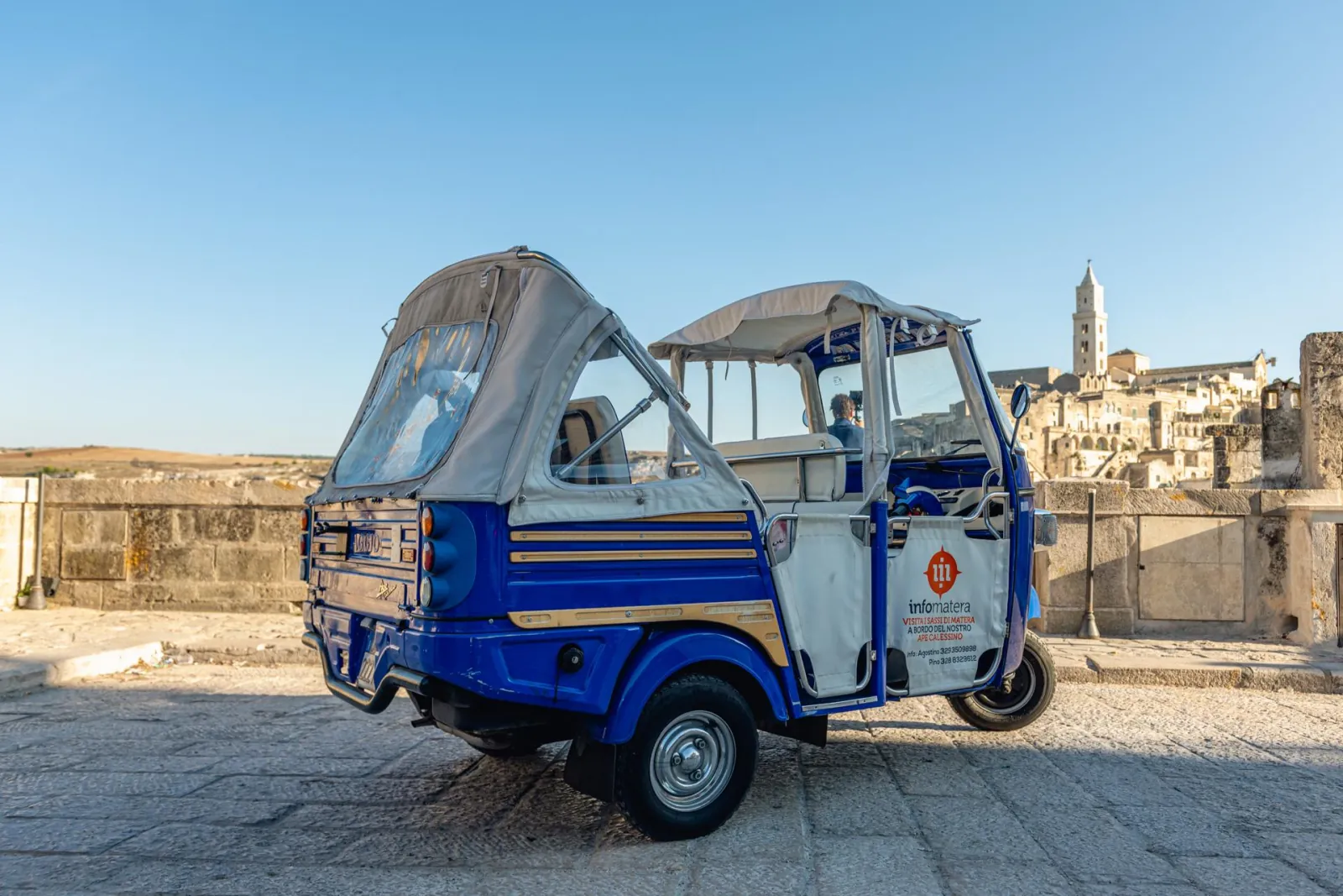 Two blue and white open-sided Ape tuk-tuks parked on a stone terrace at sunset, Matera in the distance.