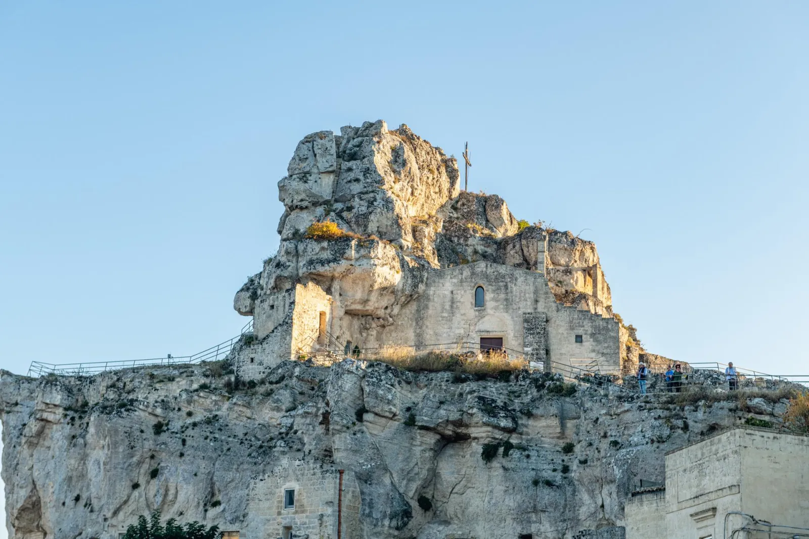A rocky crag crowned by a small rock-cut church and cross, seen against a clear sky in Matera.