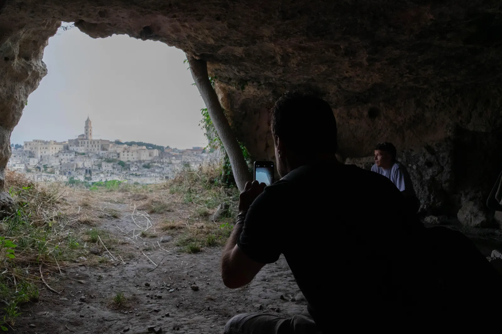 A visitor photographs the distant Matera skyline through the opening of a dark rock cave in the Murgia.