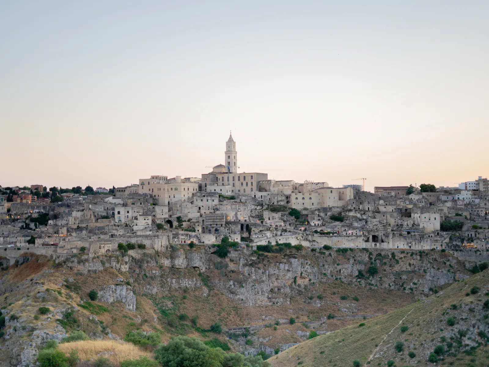 Matera's old town and cathedral tower seen across the gorge from the Murgia plateau at dusk.