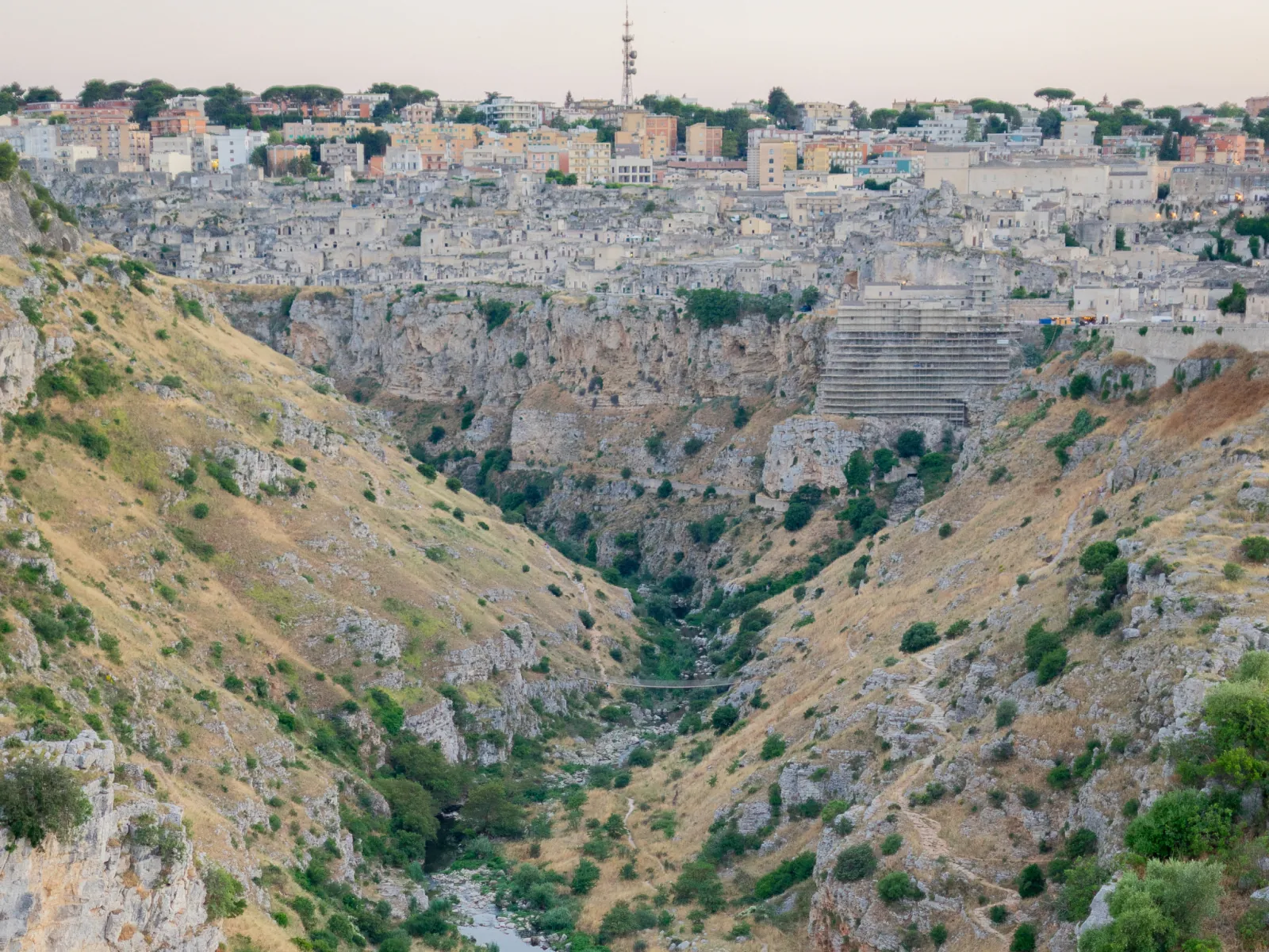 A deep, green-streaked gorge of the Gravina below Matera at dusk, with the old town on the far side.