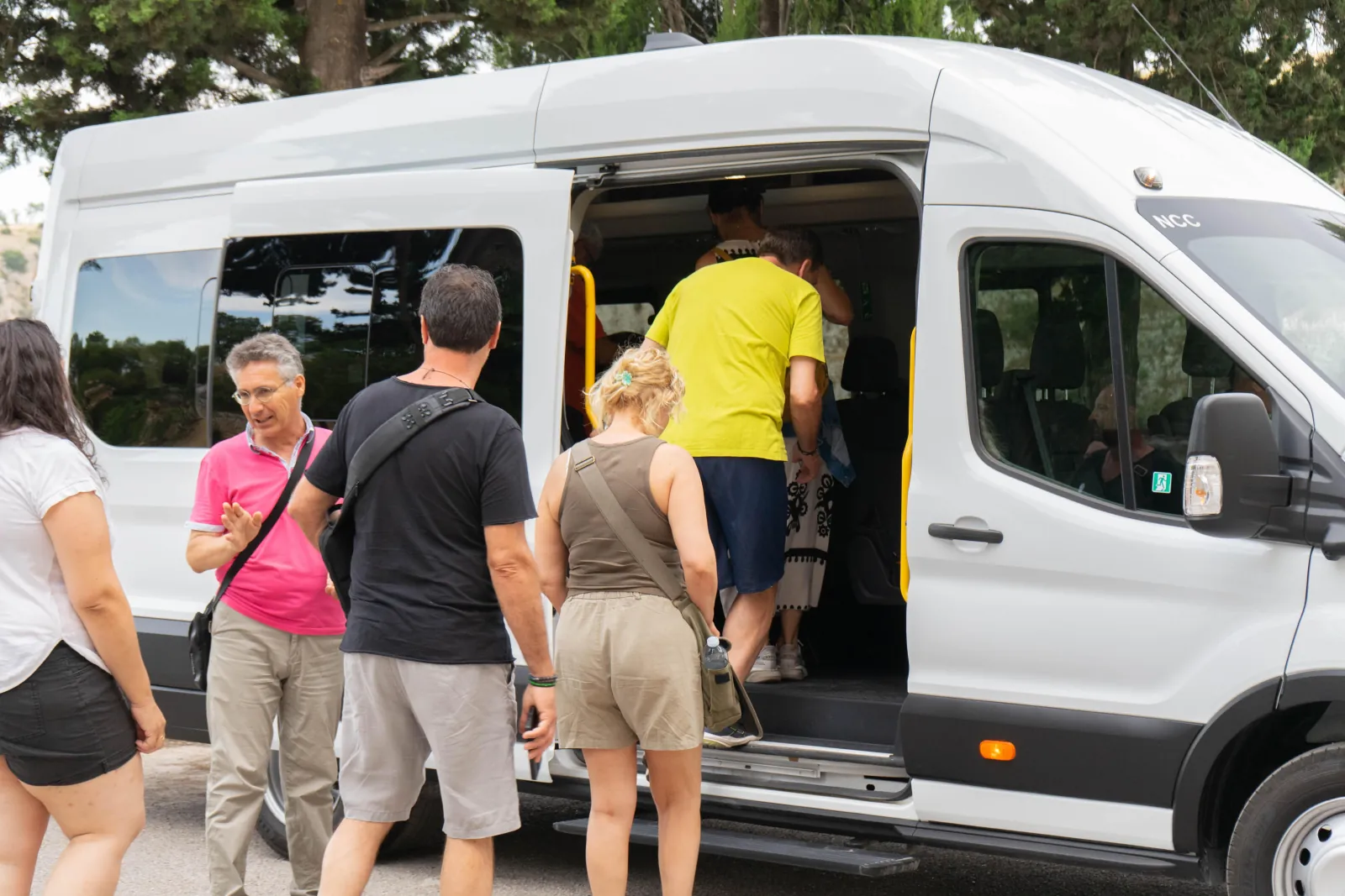 Visitors board a white tour minibus parked among trees before an excursion near Matera.